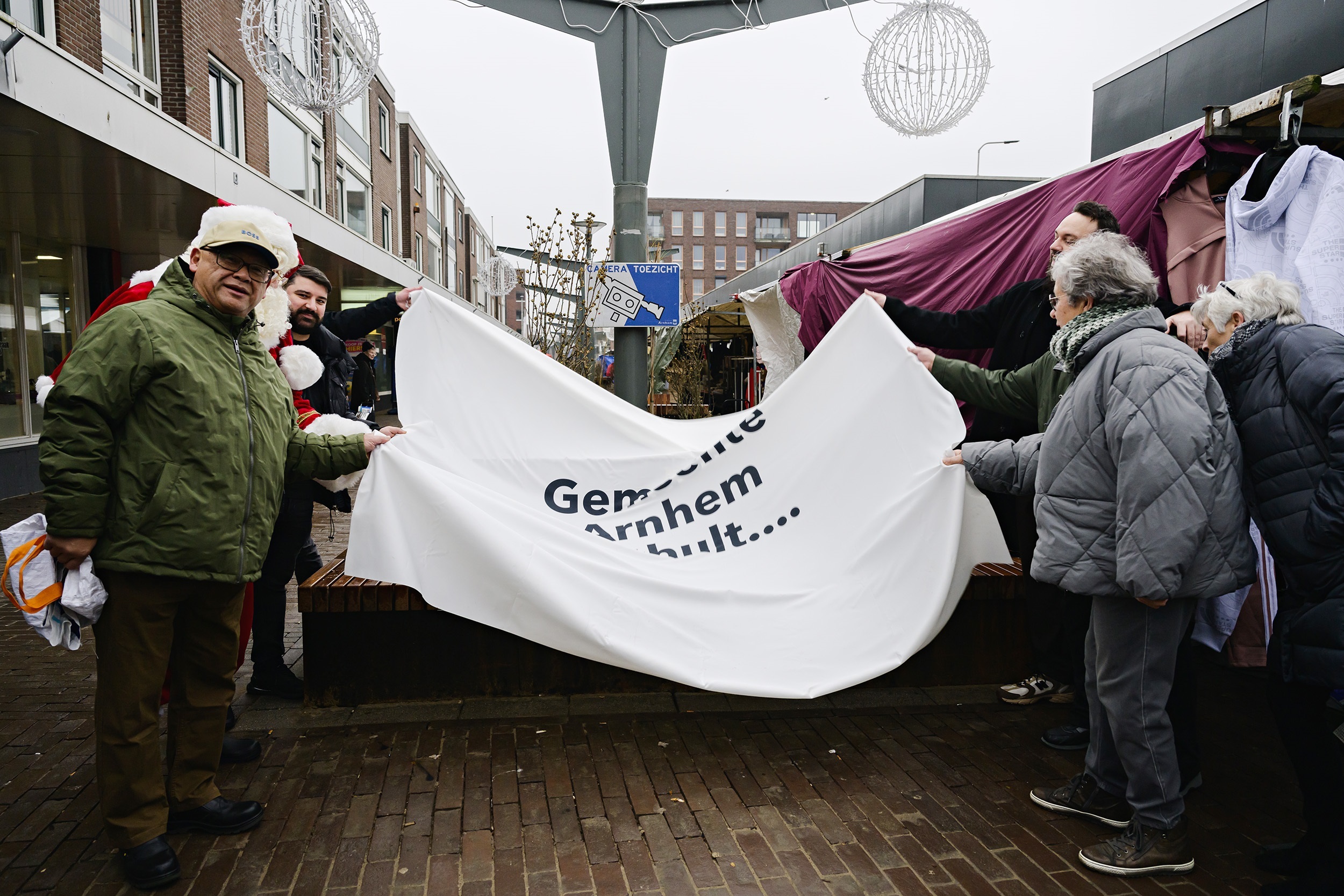 Bewoners en ondernemers van winkelcentrum Drieslag houden doek vast met de tekst 'Gemeente Arnhem onthult'.